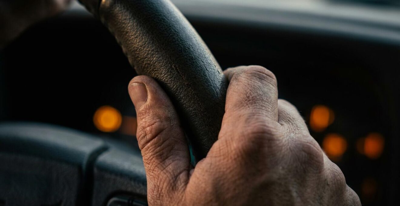 Nahaufnahme von Händen, die ein Lenkrad umgreifen, während im unscharfen Hintergrund Warnleuchten aufleuchten.